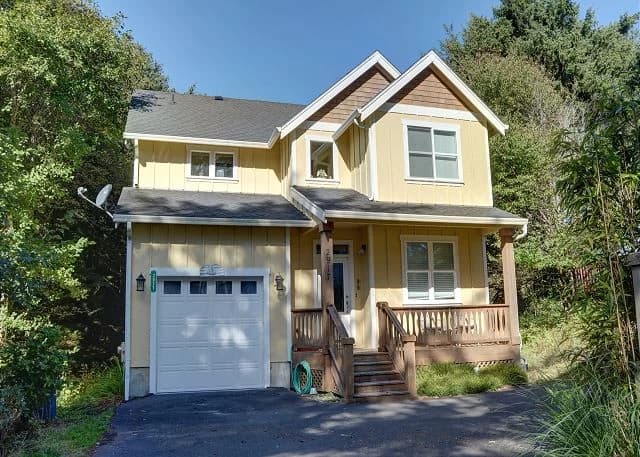 Yellow two-story house in Lincoln City with a white garage, wood porch, and green trees