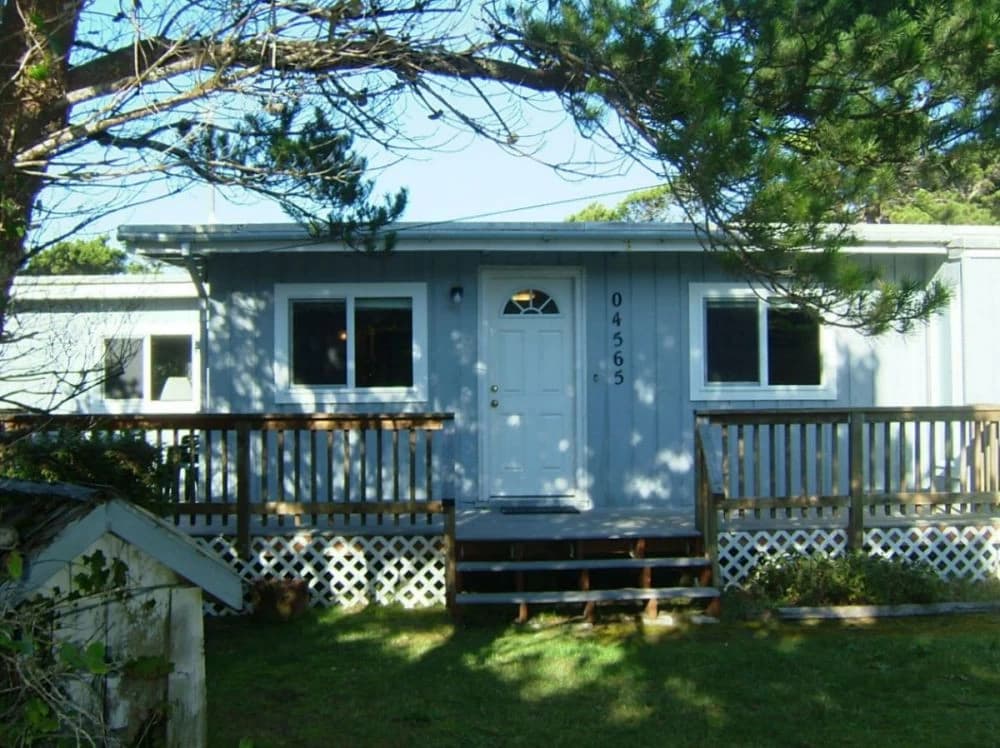 Light-blue cottage in Florence with a white door, wooden deck, and pine-shaded lawn