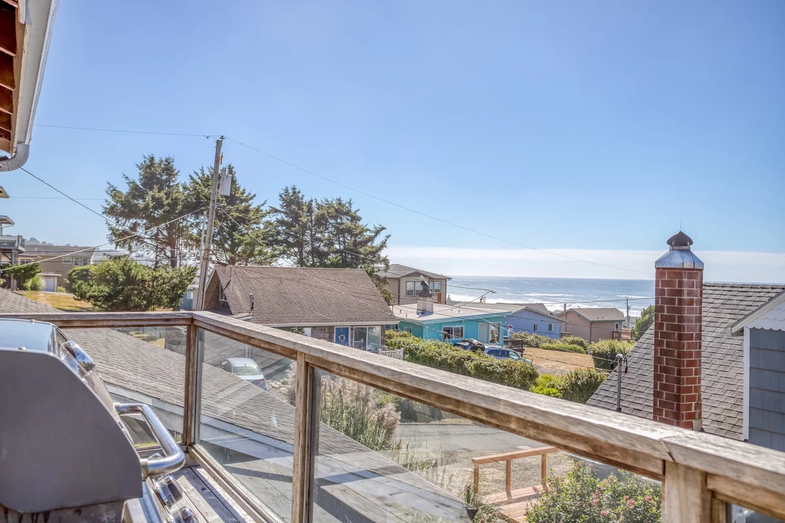 Wooden deck in Lincoln City with a glass railing, grill, and ocean beyond rooftops