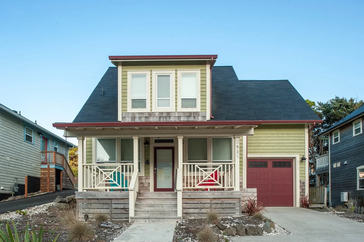 Sage-green two-story house in Lincoln City with a red door, garage, and porch chairs
