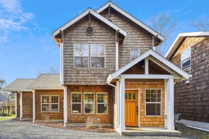 Cedar-shake Cannon Beach house with white porch pillars, a wood door, and a gravel lot