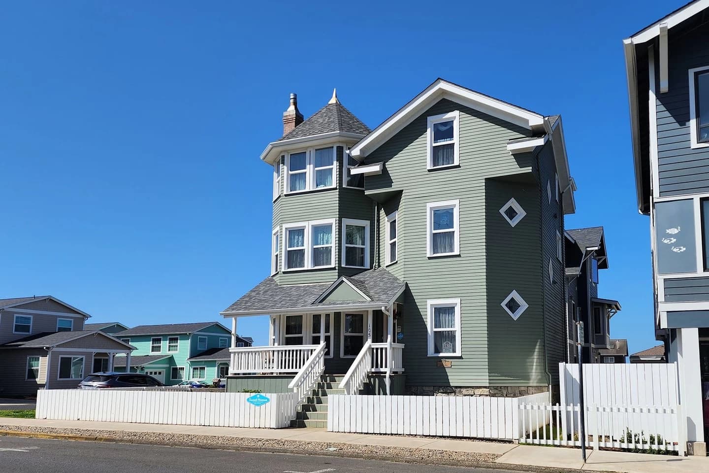 Sage-green Newport house with a turret, white picket fence, and diamond windows on the gable